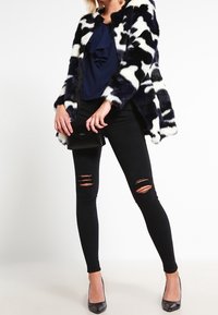 Kabát z umělé kožešiny s námořnickým a bílým vzorem, volánový námořnický top, černé slim džíny s prodřenými koleny a černé špičaté podpatky.