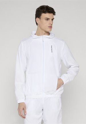 Ung mand iført en hvid hættetrøje med lynlås og hænderne i lommerne, stående mod en ensartet lys baggrund.