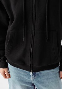 Sudadera con cremallera negra que presenta una capucha forrada, dos bolsillos frontales y puños y dobladillo acanalados. La tela parece suave y texturizada.