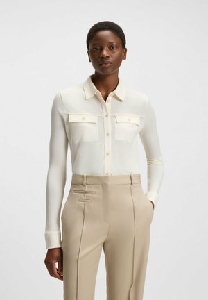 White button-up shirt with two chest pockets, long sleeves, and a smooth texture, paired with beige tailored pants featuring side accents.