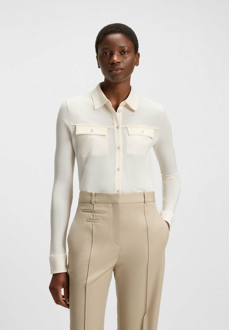 White button-up shirt with two chest pockets, long sleeves, and a smooth texture, paired with beige tailored pants featuring side accents.