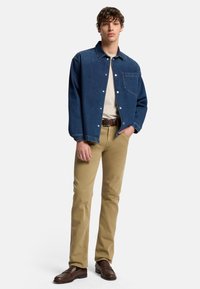 Giubbotto di denim blu scuro con bottoni a pressione, indossato sopra una camicia beige chiara. I pantaloni khaki e le scarpe marrone scuro completano l'outfit.