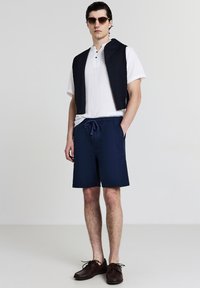Pantaloncini blu navy con vita con coulisse, abbinati a una camicia bianca con colletto e un gilet blu navy senza maniche; scarpe marroni. Tessuto dalla texture liscia.