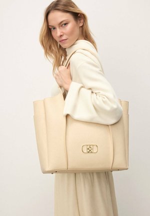 Lyshåret kvinde iført en cremefarvet dragt holder en stor beige tote-taske med guldfarvet logoemblem foran mod en ensartet lys baggrund.