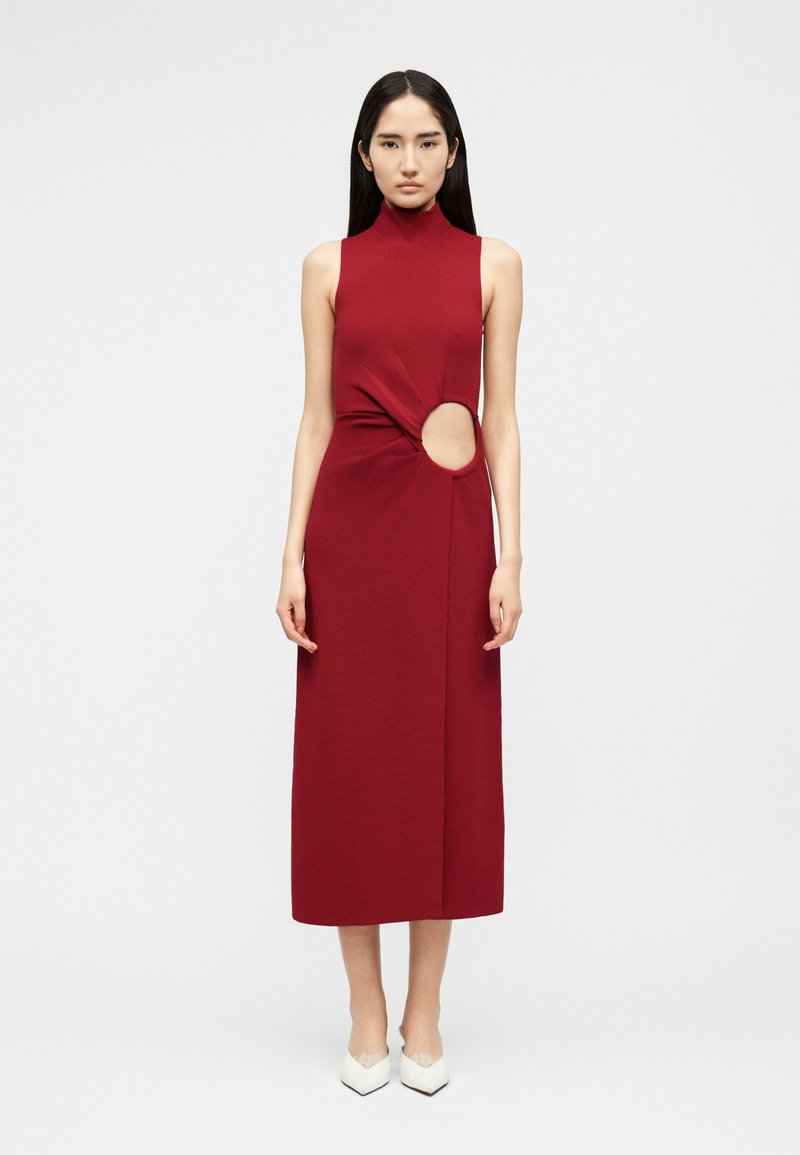 Hosszú sötét hajú nő ujjatlan, magas nyakú, piros midi ruhát visel, amelynek jobb oldalán kör alakú kivágás található deréknál, fehér hegyes orrú magassarkúval párosítva.