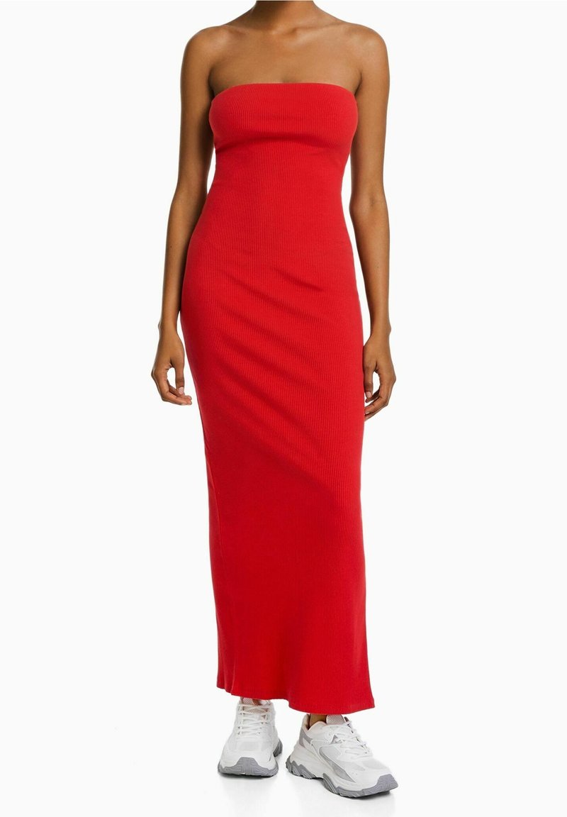 Femme portant une robe maxi rouge bustier et ajustée, associée à des baskets blanches épaisses, debout devant un fond blanc uni.