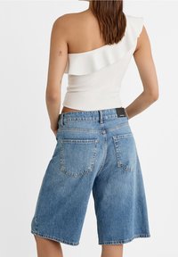 Witte top met één schouder en ruches, gecombineerd met blauwe denim shorts. De shorts hebben een relaxte pasvorm en twee achterzakken.