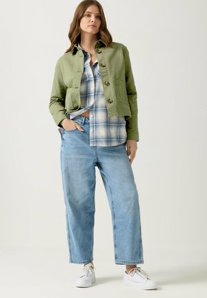 Donna che indossa jeans larghi a gamba larga di colore azzurro chiaro, sneakers bianche, una camicia a quadri blu e una giacca verde oliva con bottoni, in piedi con una mano in tasca.