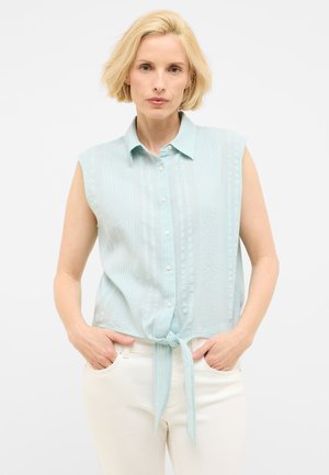 Blusa senza maniche blu chiaro con righe verticali, chiusura con bottoni e nodo sul davanti. Abbinata a jeans bianchi, che mostrano una vestibilità comoda.