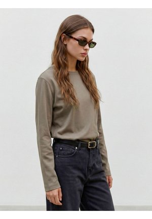 Femme aux longs cheveux ondulés portant des lunettes de soleil, une chemise taupe à manches longues rentrée dans un jean noir avec une ceinture noire, debout devant un fond uni.