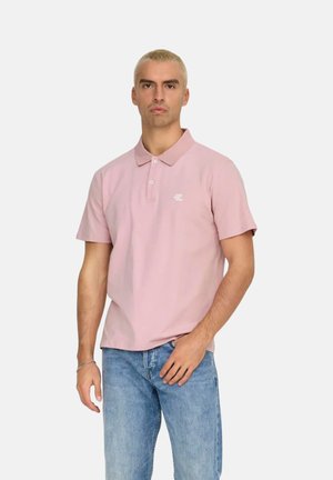 Camisa polo rosa de algodón, con cuello clásico, mangas cortas y un logo sutil en el lado izquierdo. Combinada con vaqueros azul claro.