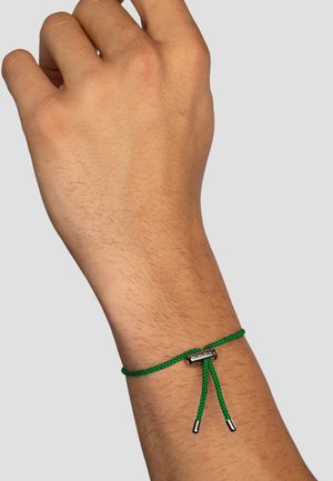 Green braided bracelet made of nylon, featuring a metal clasp and two hanging ends, worn on a hand with textured skin.