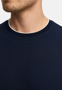 Camiseta de cuello redondo en azul marino con un tejido texturizado, que presenta un acabado en el cuello de un color claro que contrasta. Primer plano de la zona del cuello.