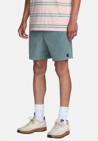 Shorts vert clair en tissu doux, avec une coupe décontractée et une taille élastique. Portés avec une chemise rayée rose et bleue et des baskets.