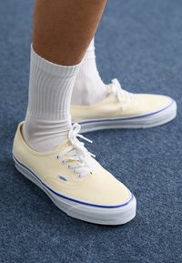 Vans sneakers in soft yellow canvas with white laces, featuring a blue stripe on the sole, paired with textured white crew socks.
