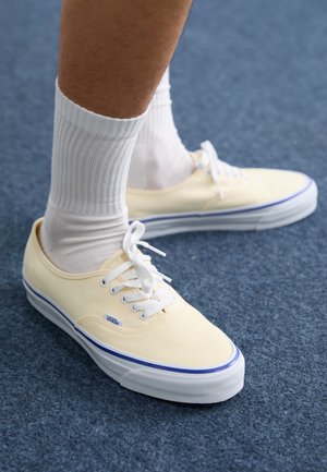 Tenisky Vans v mäkkom žltom plátne s bielymi šnúrkami, so modrým pruhom na podrážke, skombinované s textúrovanými bielymi ponožkami.