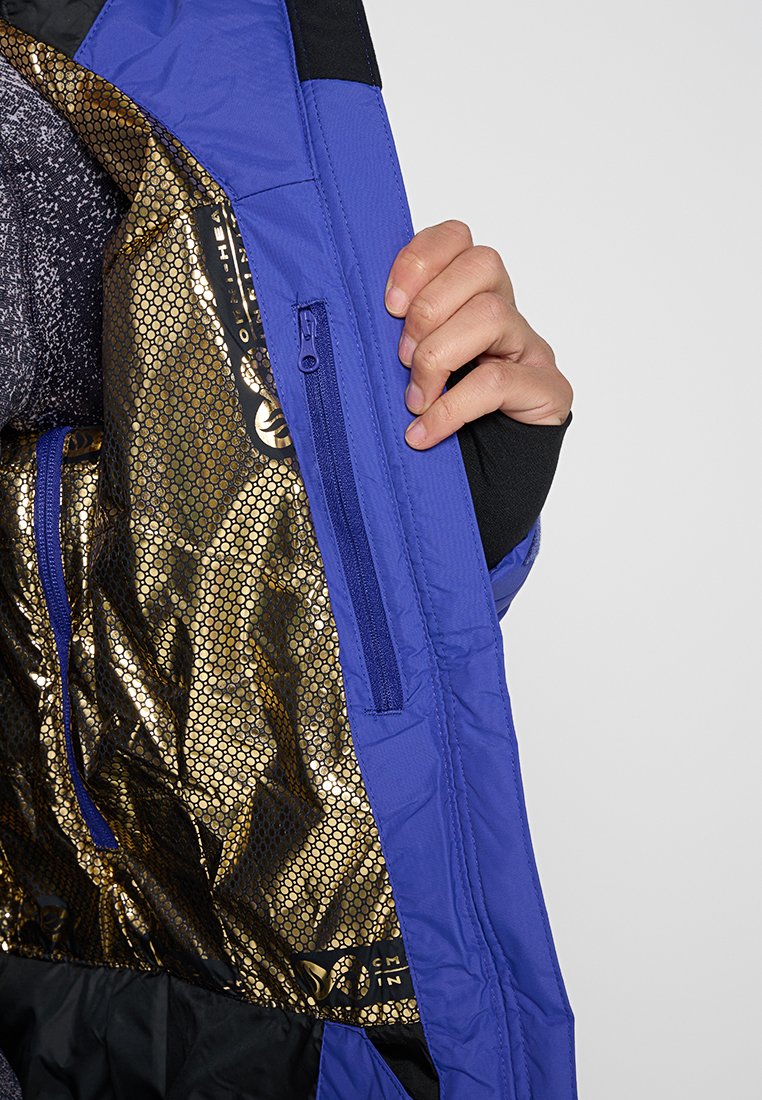 Main tenant de jaquette bleue ouverte montrant un doublure intérieure en motif de nid d'abeille doré et une poche zippée intérieure.