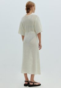 Knielanges, cremefarbenes, offenes Strickkleid mit kurzen Ärmeln und lockerer Passform, das ein strukturiertes Netzmuster und einen geraden Saum aufweist.