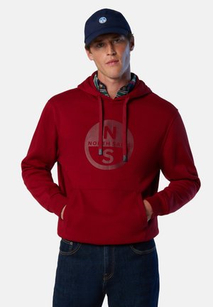 Sweat à capuche rouge avec une poche avant, un cordon de serrage ajustable et un logo circulaire comportant les lettres "N" et "S". Porté avec une chemise à carreaux et un jean bleu.