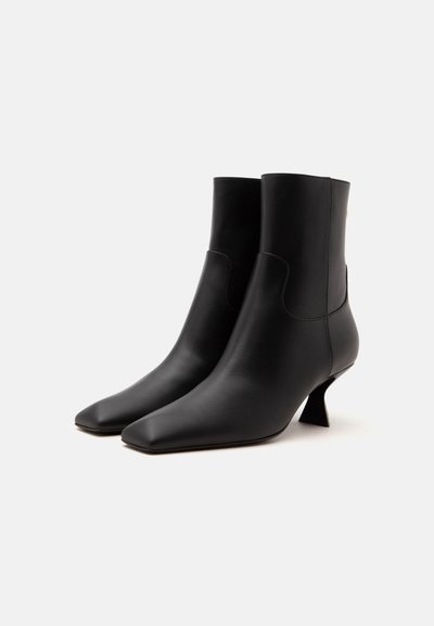 Paire de bottines en cuir noir avec bouts pointus et talons fins à la forme unique sur fond blanc.