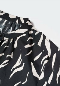 Chemise noire avec un col présentant un motif abstrait blanc. Fabriquée en tissu lisse, elle dispose d'une fermeture à boutons et de détails froncés.