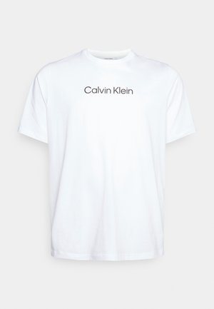 Bílé tričko s krátkým rukávem s nápisem "Calvin Klein" uprostřed na hrudi na jednoduchém světlejším pozadí.