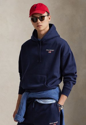 Μπλε σκούρο φούτερ με κουκούλα, λογότυπο "POLO SPORT", κορδόνια και τσέπη καγκουρό. Φορεμένο με μπλε πλεκτό πουλόβερ δεμένο στη μέση, κόκκινο καπέλο και γυαλιά ηλίου.