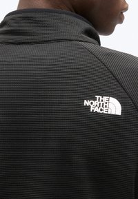 Μαύρο υφασμάτινο μπουφάν που φοράει άτομο, με το λογότυπο The North Face σε λευκό στη δεξιά πλευρά του πάνω μέρους της πλάτης.