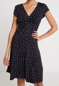 Robe noire à pois blancs, manches courtes, décolleté en V, détail noué au niveau de la poitrine, jupe évasée, texture de tissu lisse.