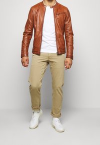 Brun läderjacka med en dragkedjeficka, vit t-shirt och beige byxor. Modell som bär vita sneakers och står mot en neutral bakgrund.