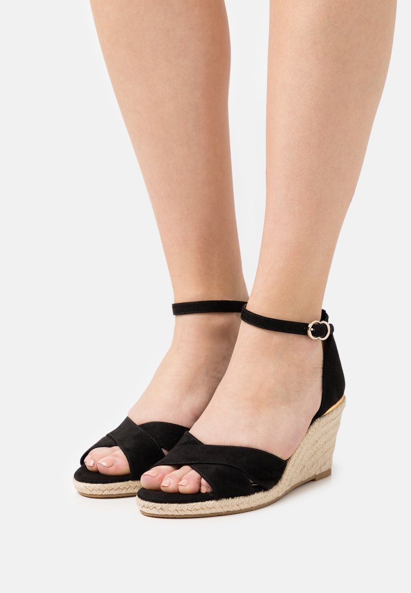 Pieds portant des sandales compensées en daim noir avec des brides à la cheville et des talons compensés en jute tressée sur un fond clair uni.