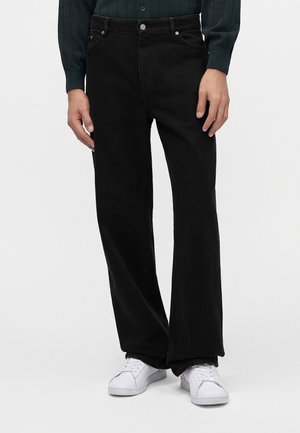 Personne portant un jean noir ample avec des poches devant et des baskets blanches, debout devant un fond clair uni.