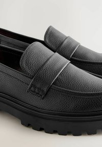 Mocasines de cuero negro con un acabado texturizado, un perfil plano y una suela de goma con patrón. Presenta una correa prominente a lo largo del empeine.