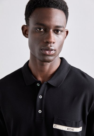 Svart polo skjorta med korta ärmar, krage, tre knappar och en bröstficka med en vit rand och "MOSCHINO" etikett. Slät textur.