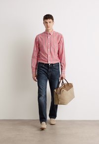 Camisa de botones a rayas rojas y blancas, jeans oscuros, bolsa beige con acentos marrones, zapatos claros. Modelo caminando sobre una superficie neutra.
