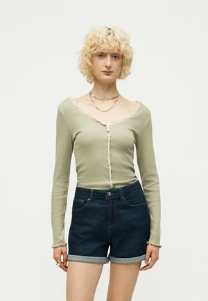 Giovane con capelli biondi ricci indossa una maglia verde chiaro a maniche lunghe con bottoni e pantaloncini di jeans scuri risvoltati, in piedi su sfondo bianco.