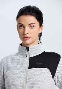 Chaqueta de forro polar en gris claro con un patrón texturizado, con un acento negro en los hombros, cuello alto y cierre de cremallera.