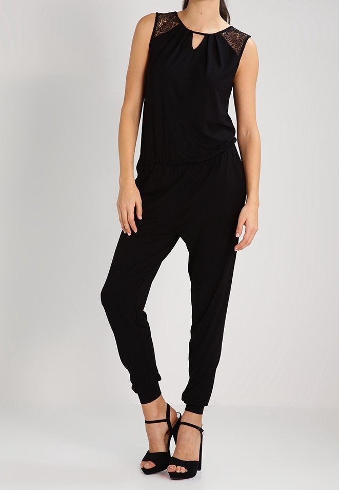 Combinaison noire sans manches avec détails en dentelle sur les épaules, encolure en trou de serrure, jambes fuselées, portée avec des sandales noires à talons blocs.