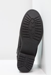 Suela de bota negra con goma texturizada y patrones de tracción, que incluyen líneas y formas para un mejor agarre. Diseño robusto con bordes reforzados.