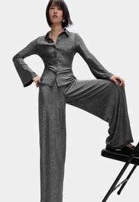 Glanzend, donkergrijs kostuum met een aansluitend overhemd en wijdvallende broek. De stof heeft een subtiele glans en een gladde textuur.