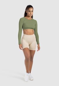Olivgrünes langärmeliges Crop-Top mit rundem Ausschnitt, kombiniert mit beigefarbenen, hoch taillierten Shorts, ergänzt durch weiße Sportschuhe.