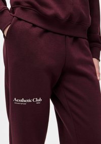 Burgundsko trenirka iz mehkega blaga z bočnim žepom in belim napisom "Aesthetic Club established 1892" na stegenu.