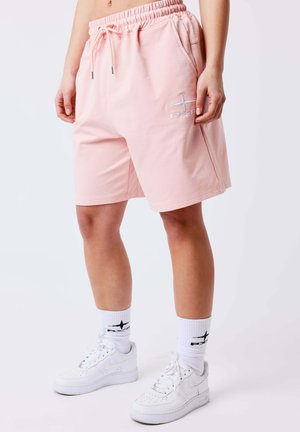 Personne portant un short rose pâle avec le logo "PXFD", des chaussettes blanches avec le même logo, et des baskets blanches Nike sur un fond blanc.