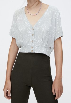 Jasnoszara bluzka z krótkim rękawem w białe kropki, z dekoltem w kształcie V, zapinana na guziki i krótka, zestawiona z ciemnozielonymi spodniami o wysokim stanie.