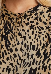 Blusa khaki con una stampa animale astratta nera e motivi a stella, chiusura con bottoni al collo e tessuto morbido e testurizzato.