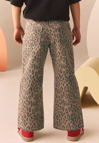 Pantaloni a zampa d'elefante in stampa leopardata beige e blu denim, con tasche posteriori e una vestibilità ampia. Abbinati a sneakers rosse su uno sfondo morbido.