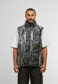 Gilet sans manches rembourré, avec un design à motifs noir et blanc, un col haut, une fermeture éclair et des poches latérales. Des accents géométriques sont présents.