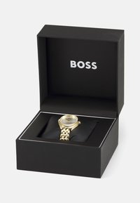 Montre en or avec un cadran rond et un bracelet argenté présentée dans une boîte noire doublée de tissu doux. La boîte arbore un logo "BOSS" en blanc.