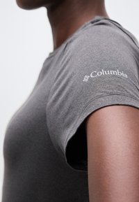 Κοντινό πλάνο ατόμου που φοράει γκρι μπλουζάκι Columbia, με το λογότυπο στο δεξί μανίκι, σε απλό φόντο.