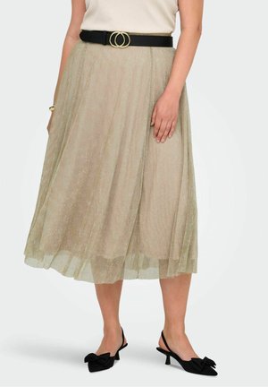 Femme portant une jupe midi beige avec une ceinture noire et des chaussures noires à talons bas avec des nœuds, debout devant un fond uni.